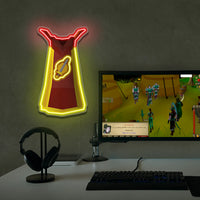 Neon sign Runescape Attack Cape of a red cape with a sword on a wall above a computer setup playing a video game.