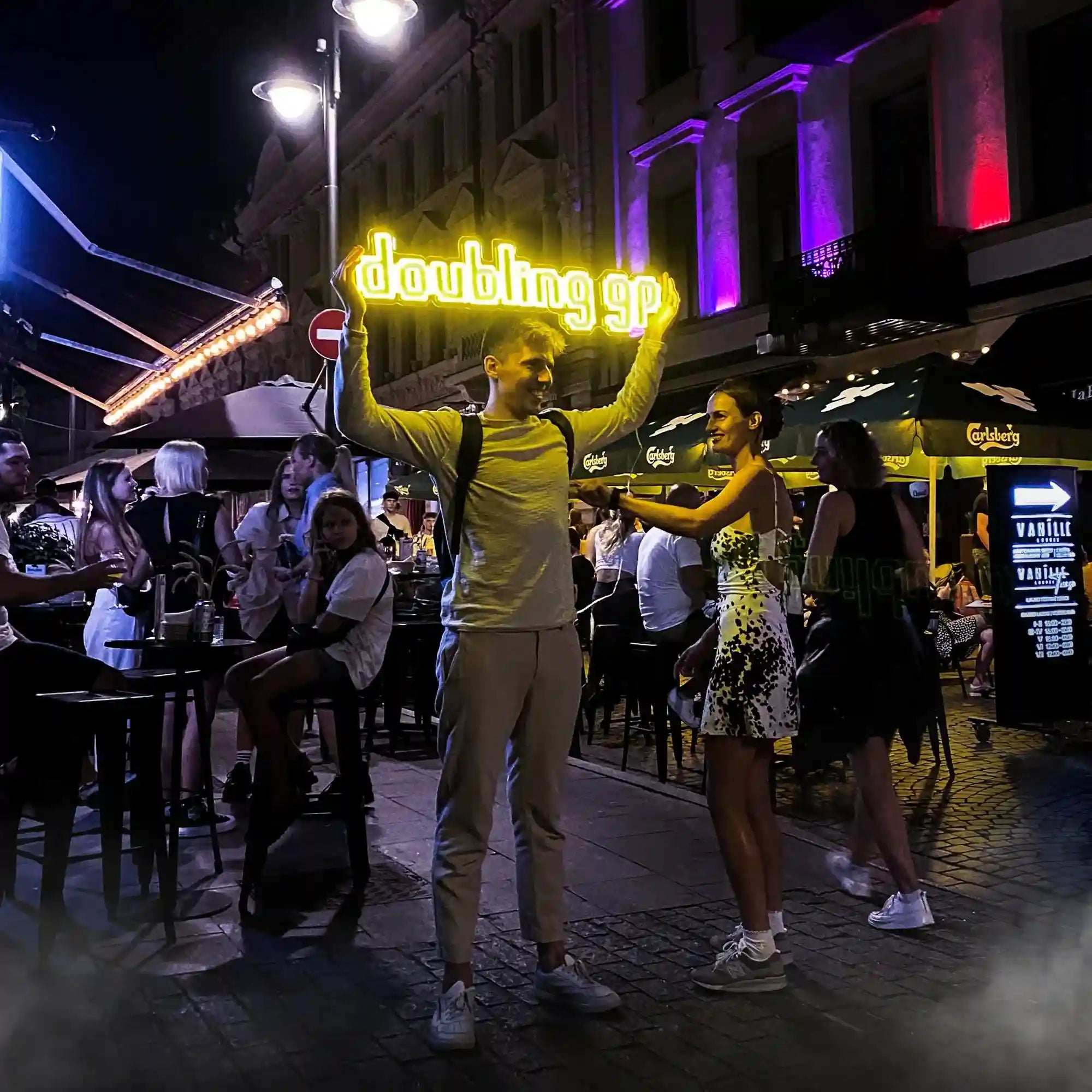 A person proudly displays the Runescape Doubling GP LED Neon Sign in a bustling city center, showcasing Runescape's iconic culture in public. Ideal for Runescape enthusiasts seeking nostalgic decor featuring keywords like Runescape, Doubling GP, custom LED neon sign, and Runescape memorabilia.